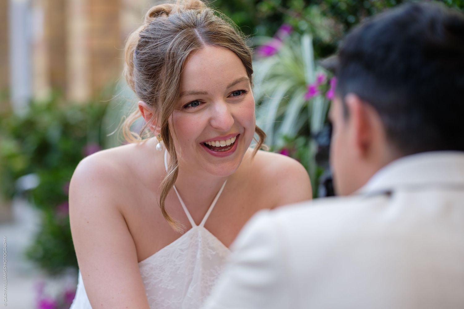 Bride in white halter dress laughing joyfully during outdoor wedding ceremony with lush greenery background.