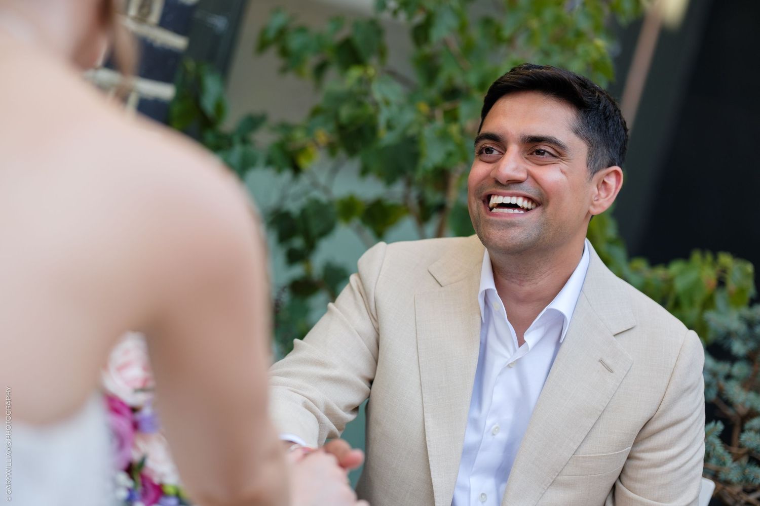 Smiling man in beige suit holding hands with woman in floral dress at outdoor wedding ceremony.