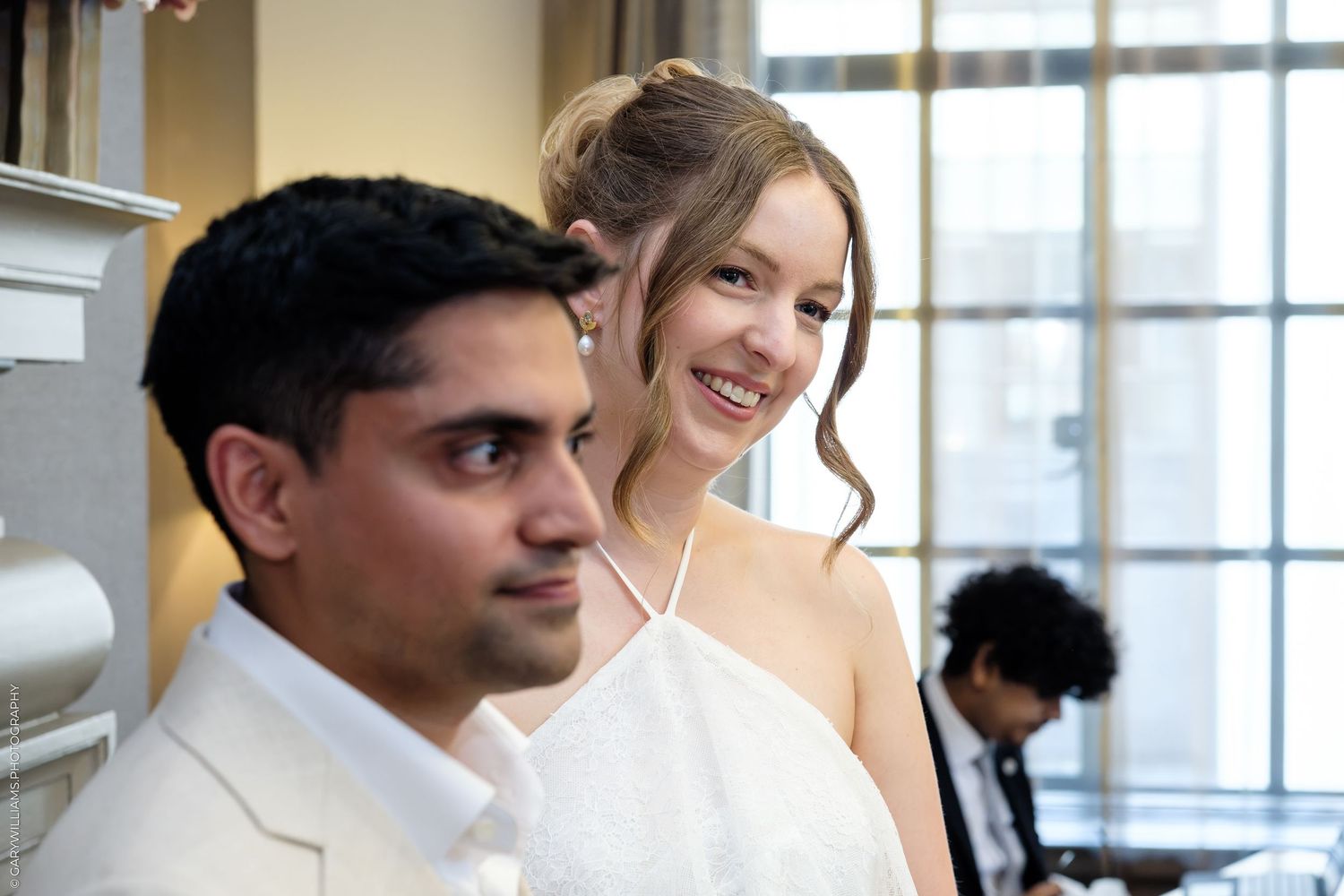 Smiling bride in white halter dress standing beside a man in a white suit at a wedding reception.