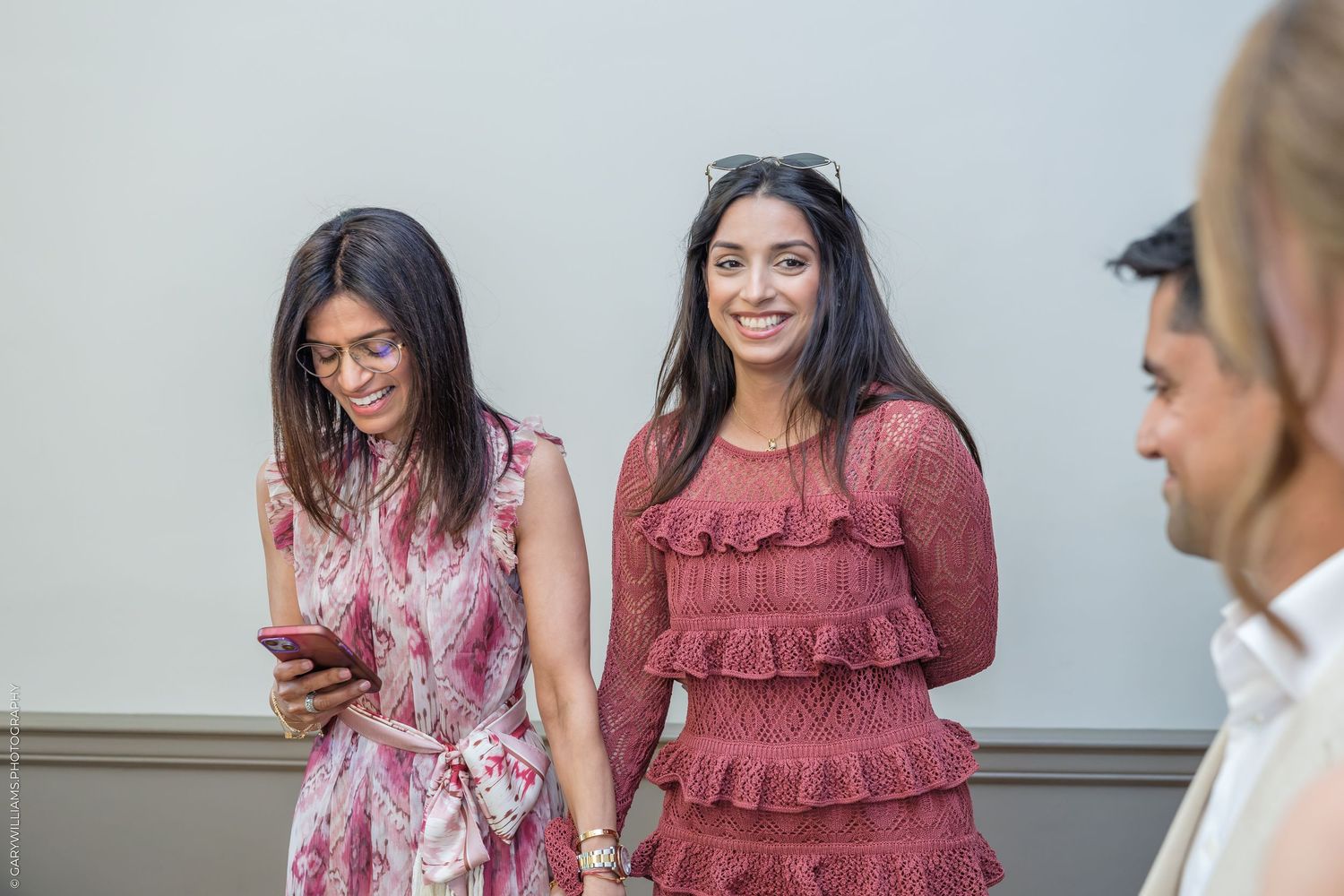 Two women in pink dresses smile outdoors, one checking her phone, with a man visible in the foreground.