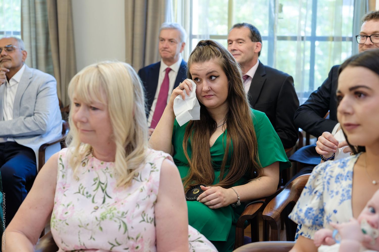 Emotional woman in green dress wiping tears at a formal indoor ceremony, surrounded by attendees in smart attire.