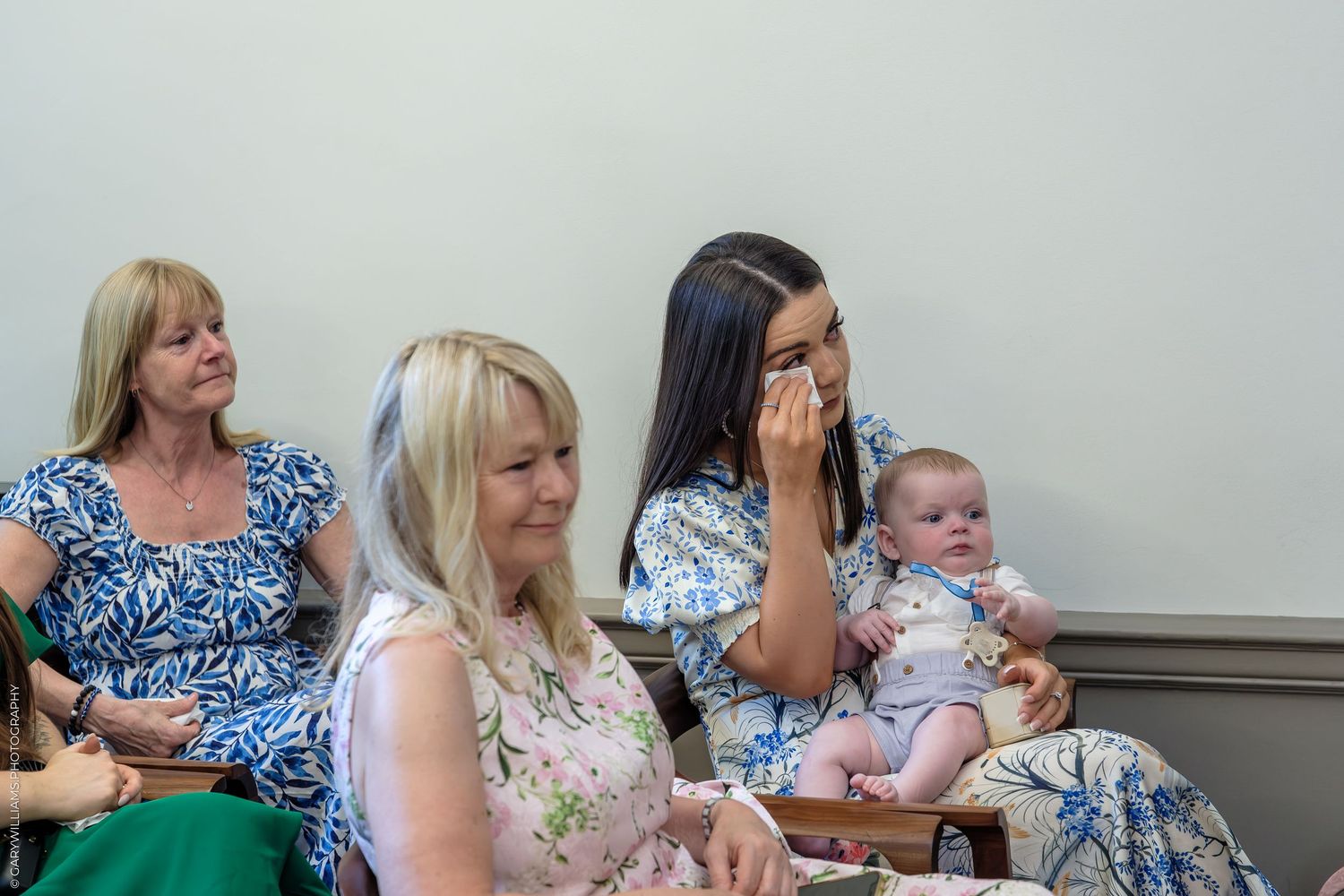 Women and baby seated at an indoor event, one wiping tears while holding an infant.