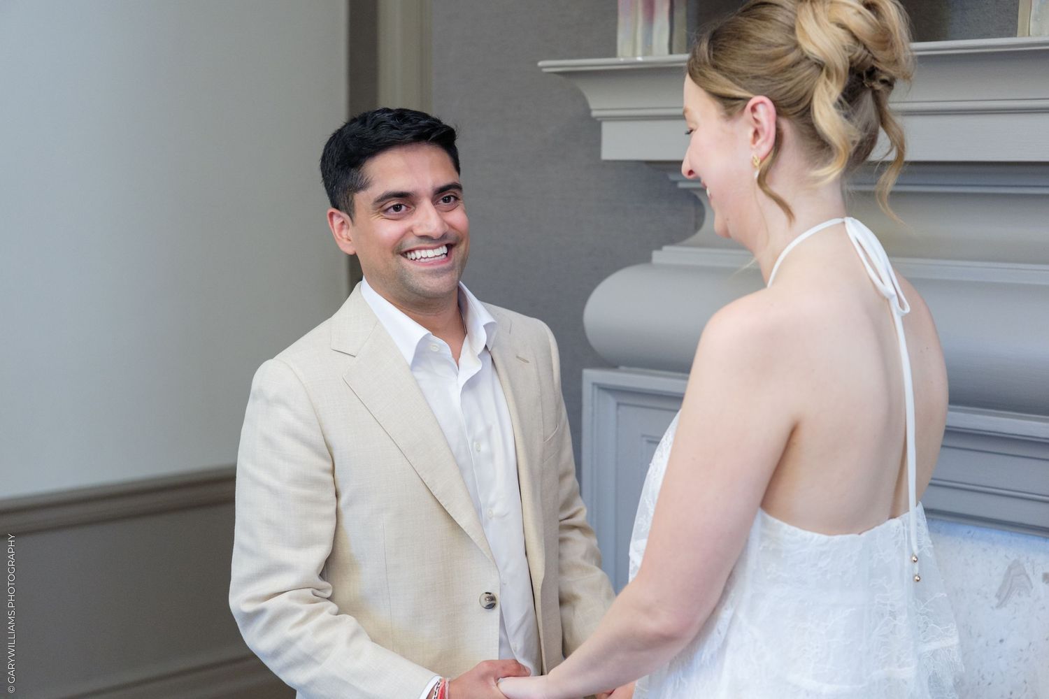 Groom in beige suit smiling at bride during wedding ceremony indoors.