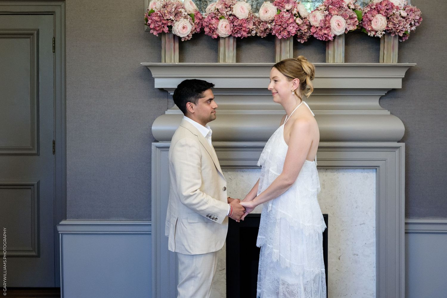 Couple holding hands at wedding ceremony before floral mantelpiece with pink and white flowers.