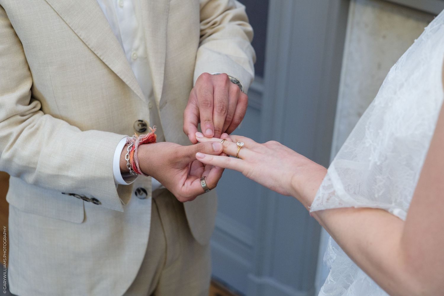 Groom placing a wedding ring on bride's finger during ceremony, groom wearing beige suit with red bracelet.