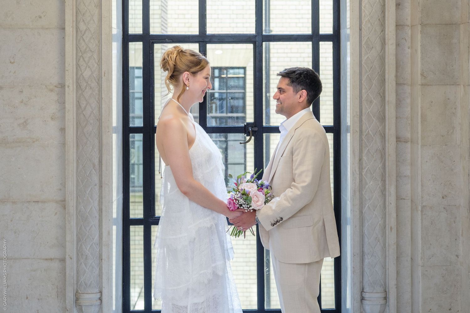 Bride and groom holding hands and exchanging vows in an elegant indoor venue with large windows.