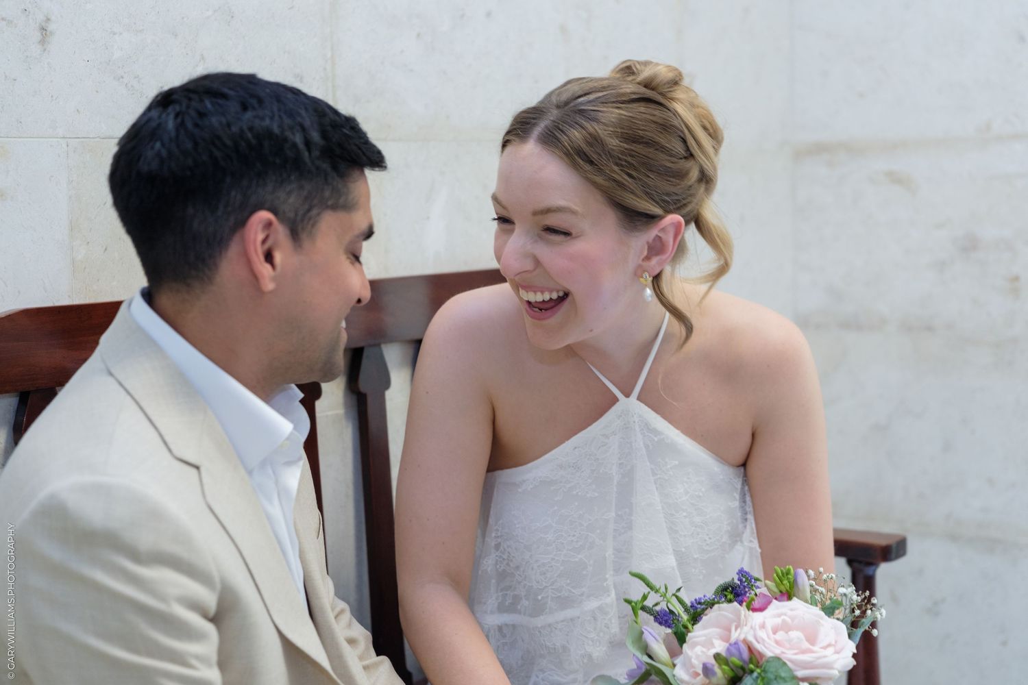 A joyful bride in a white halter gown laughs with her groom in a beige suit during their wedding ceremony.