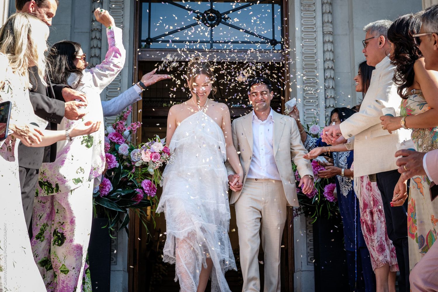 Bride and groom exit wedding venue as guests toss confetti, surrounded by flowers and celebration.