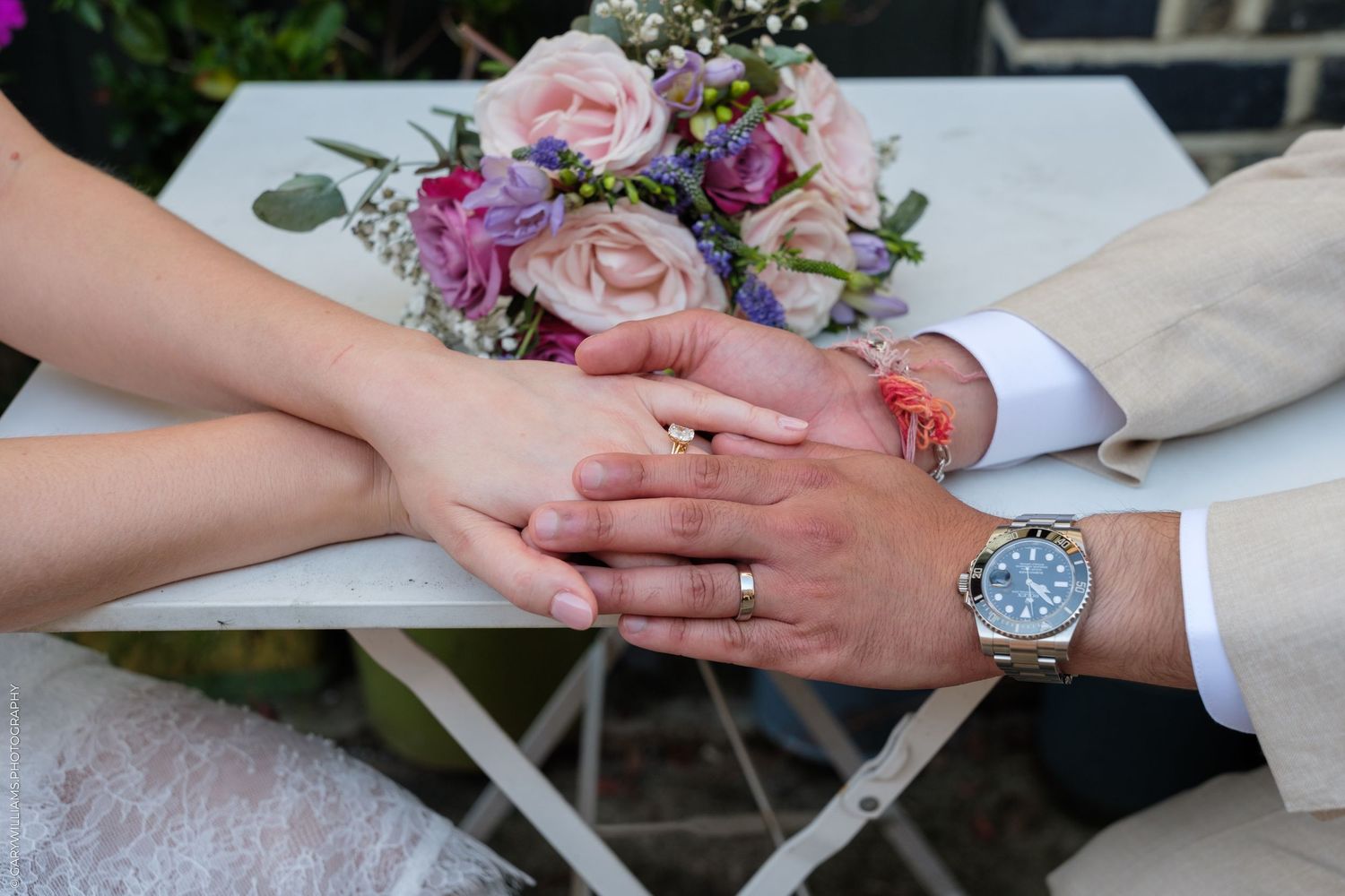 Newlyweds holding hands with wedding rings on white table beside colorful bridal bouquet.
