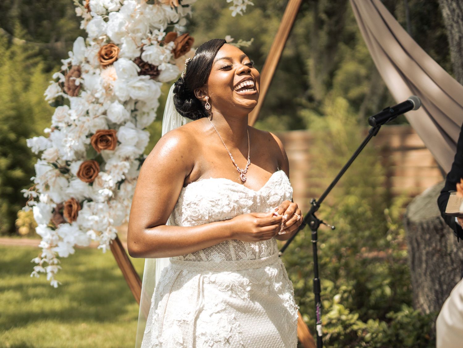 Joyful Black bride in white lace strapless gown laughing at outdoor wedding ceremony with floral arch at Idlewood Venue in Bartow