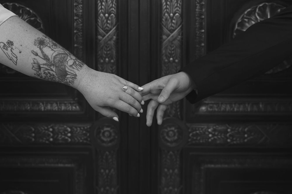 Black and white photo of two hands touching, one with a tattoo sleeve, against an ornate dark door.