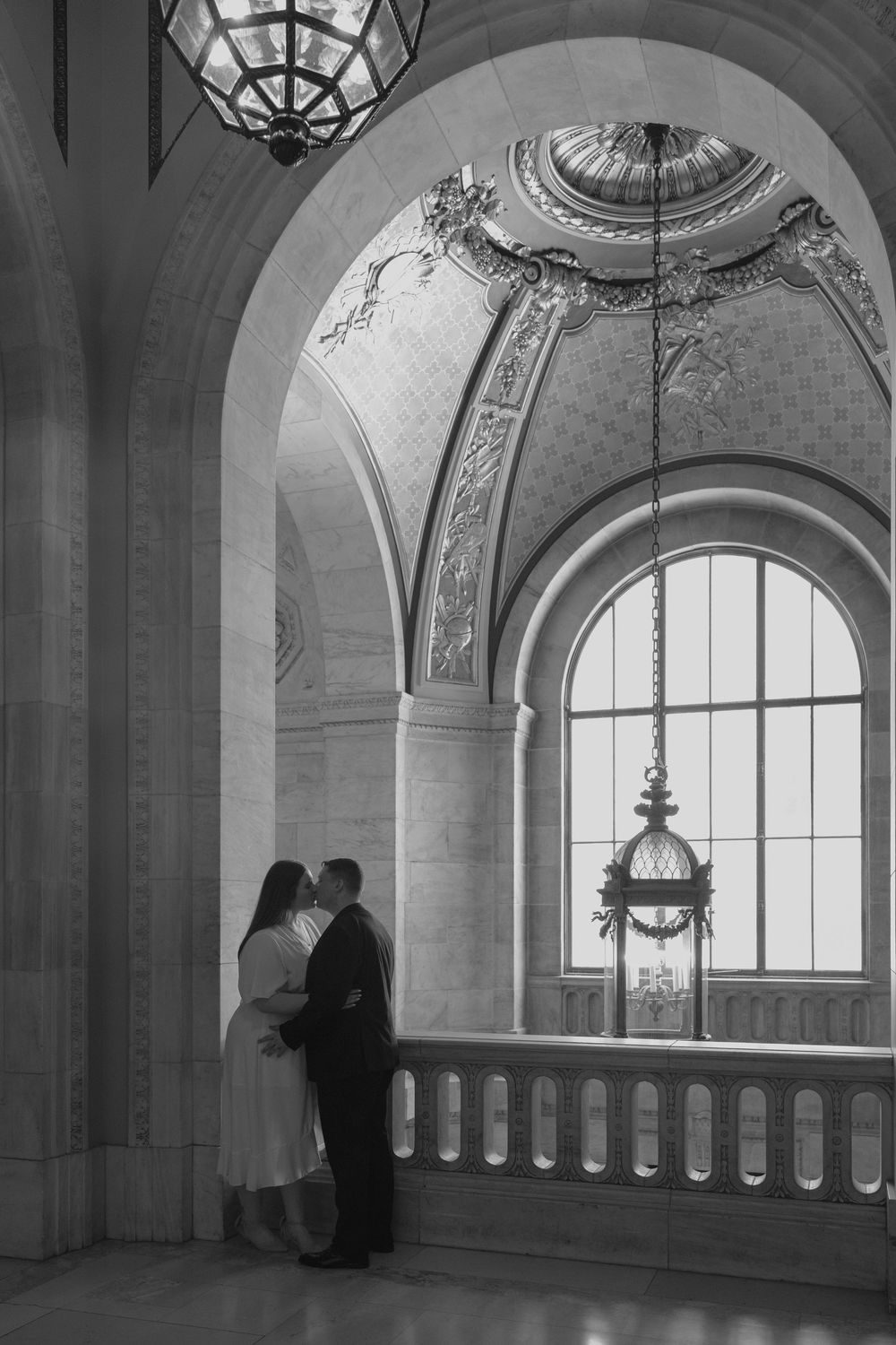 Couple sharing a romantic kiss in an ornate beaux-arts building with arched windows and decorative ceiling dome.