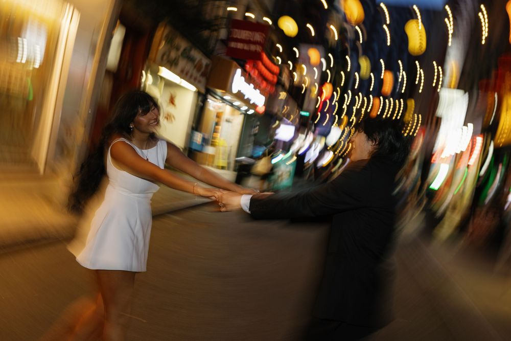 Couple holding hands dancing on a vibrant, blurred city street at night with colorful lights.