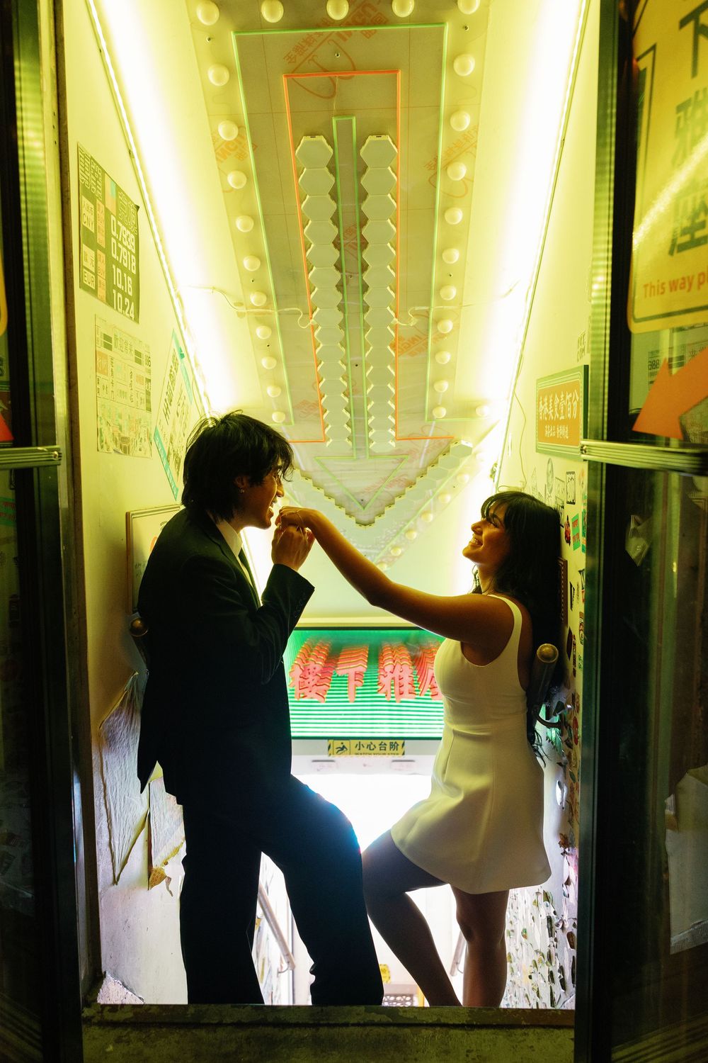 Couple sharing a romantic moment in a glowing illuminated elevator, woman in white dress touching man's face.