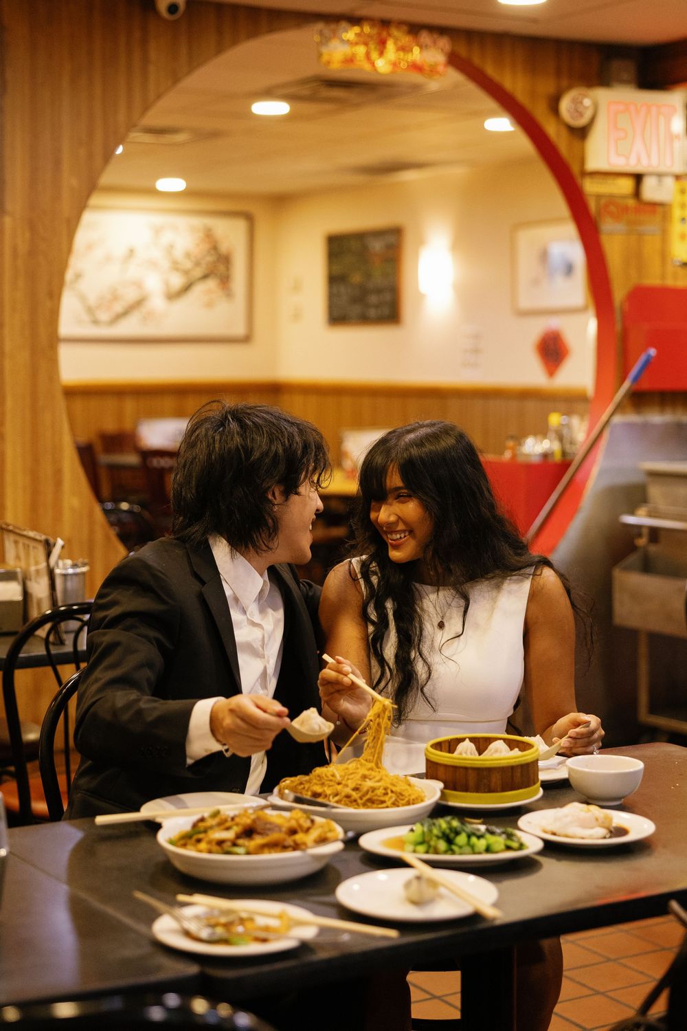 Couple sharing noodles and dim sum at a Chinese restaurant, dressed in formal attire, smiling at each other.