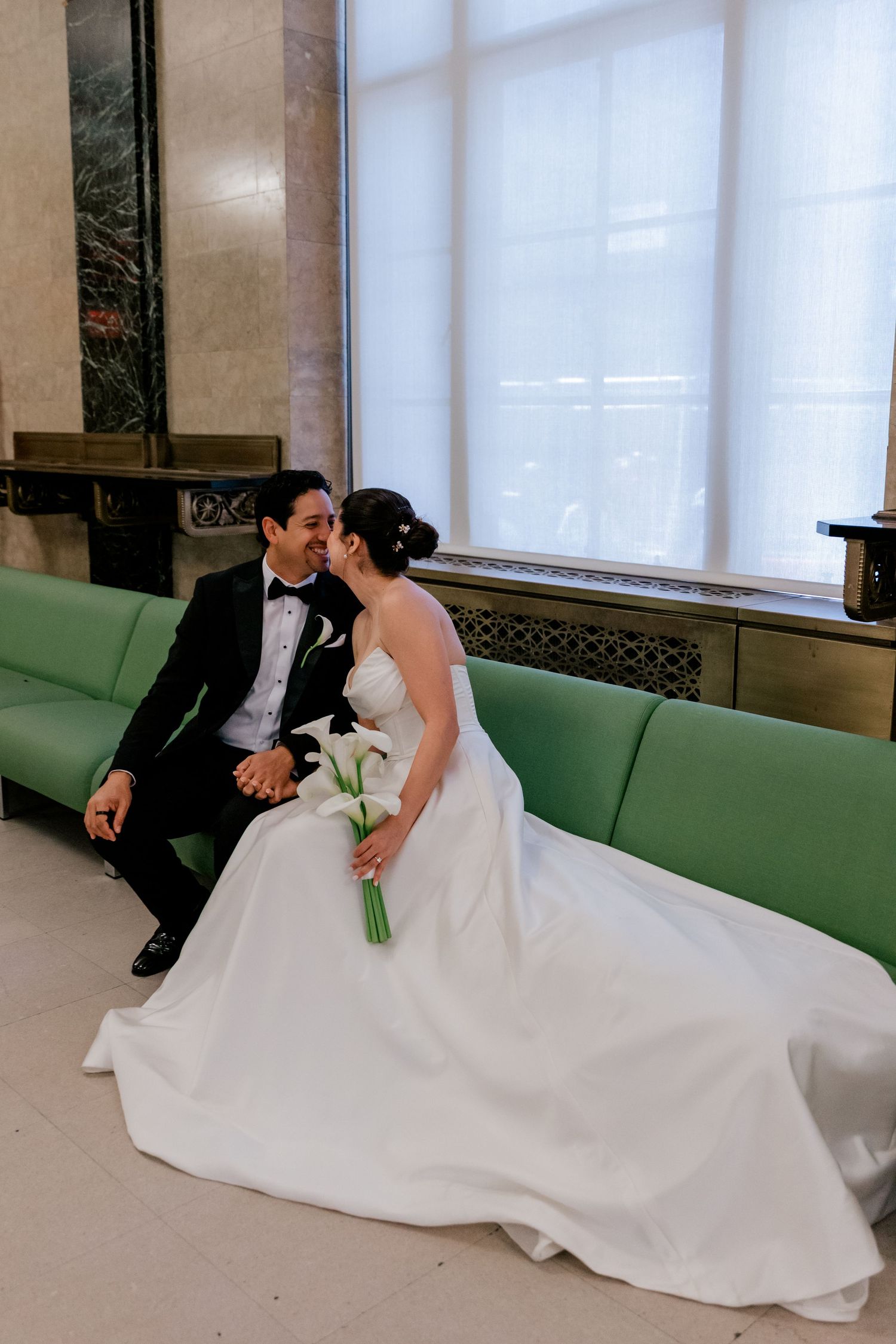 Bride and groom share a romantic moment on a green velvet bench, bride holding white calla lilies in elegant ballgown.