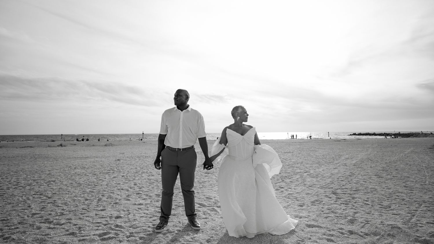 Black and white photo of a couple in wedding attire holding hands on a sandy beach with ocean horizon behind them.