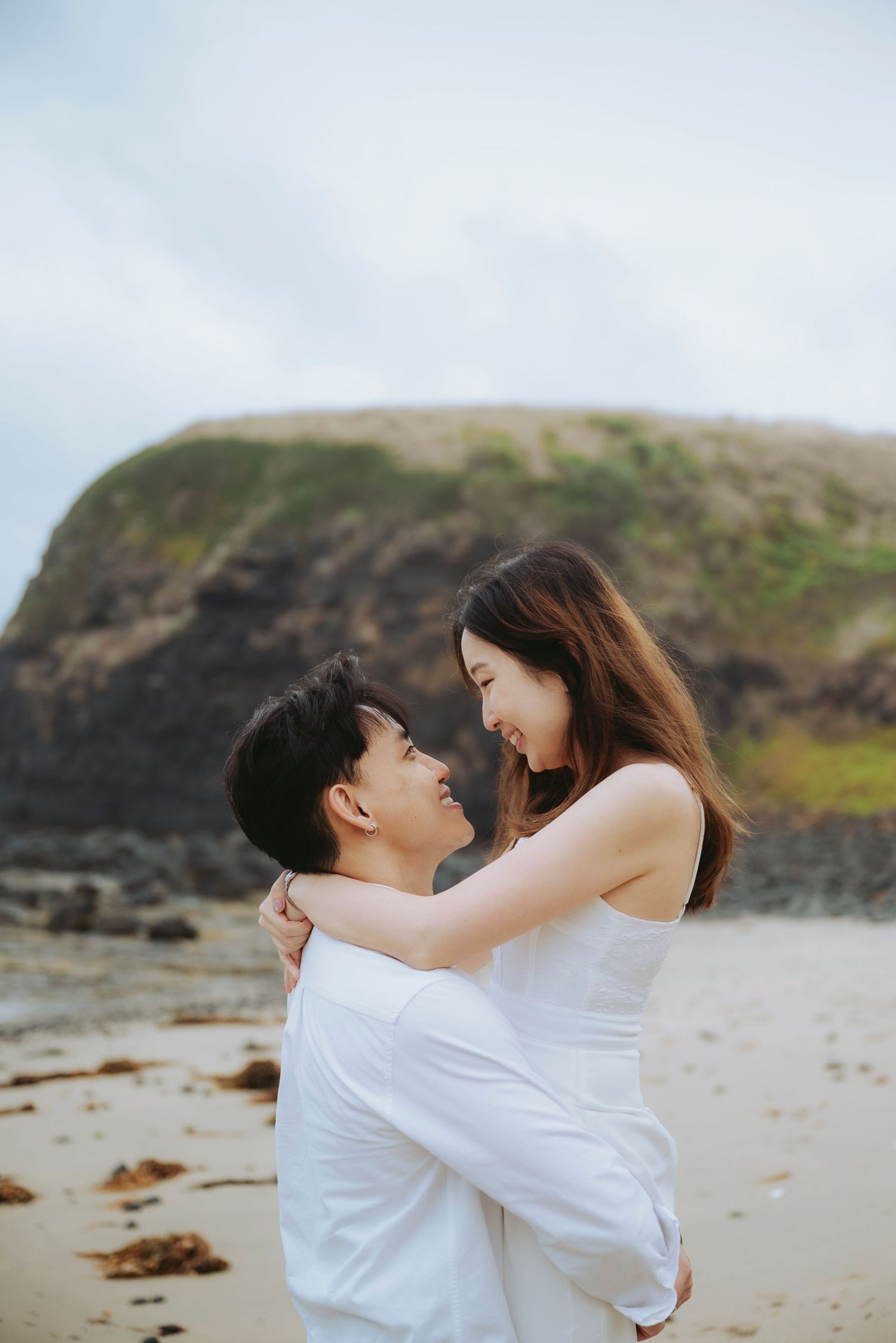 A couple embraces on a sandy beach, the man lifting the woman in a white dress with rocky cliffs in the background.