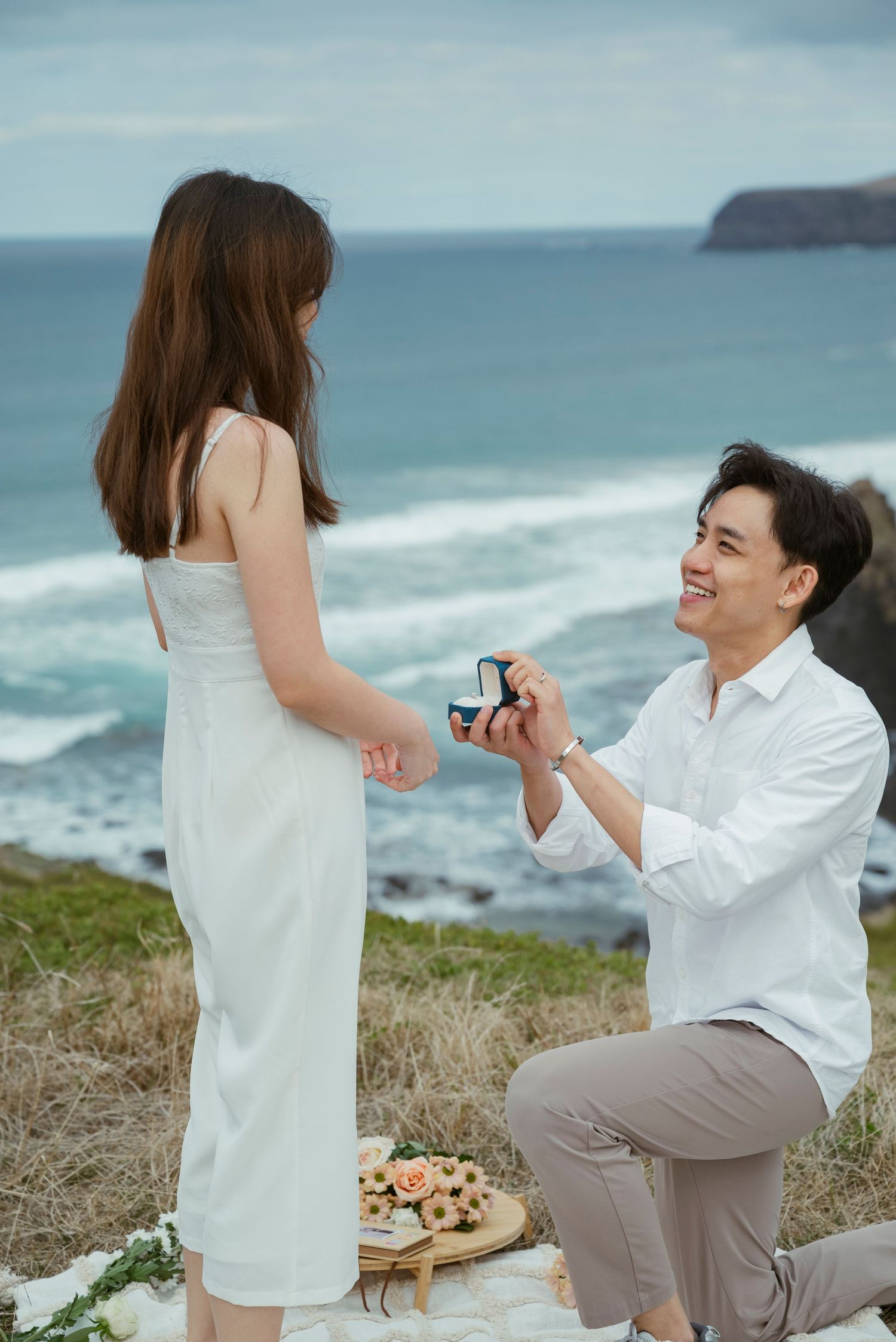 Man proposes to woman on one knee by the ocean, holding a ring box, both dressed in white and gray.