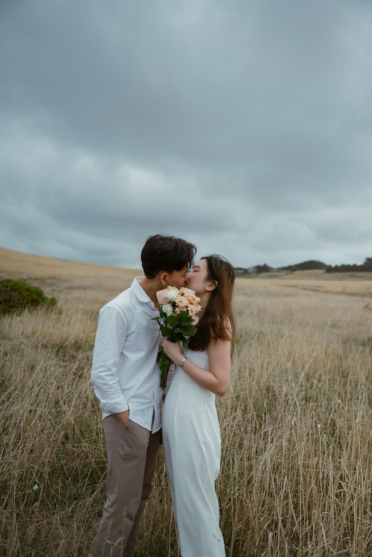 Couple kissing in golden field holding bouquet, wearing white dress and shirt, under cloudy sky.