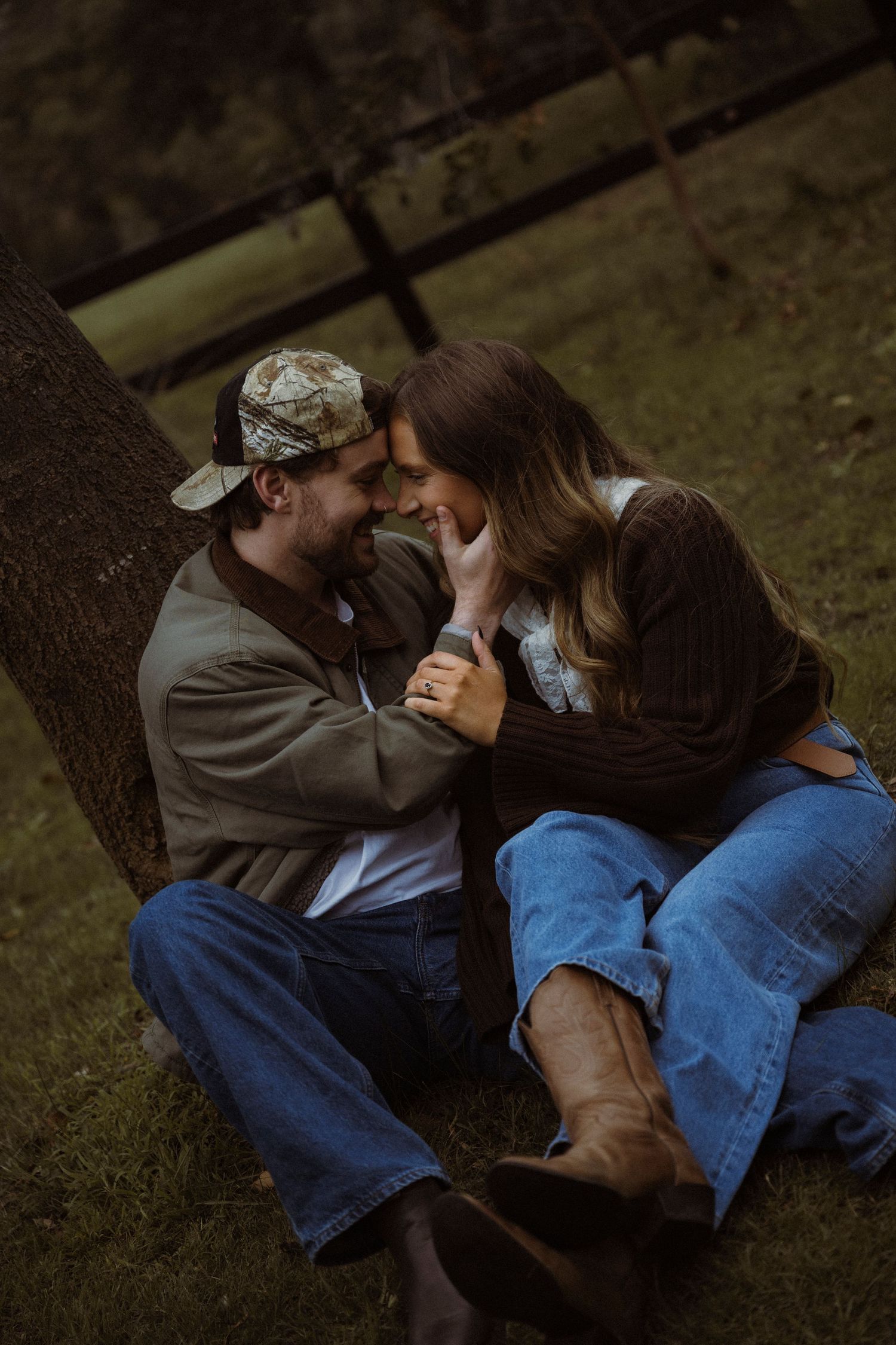 A couple sits against a tree in jeans and boots, sharing a romantic moment outdoors in a rustic countryside setting.