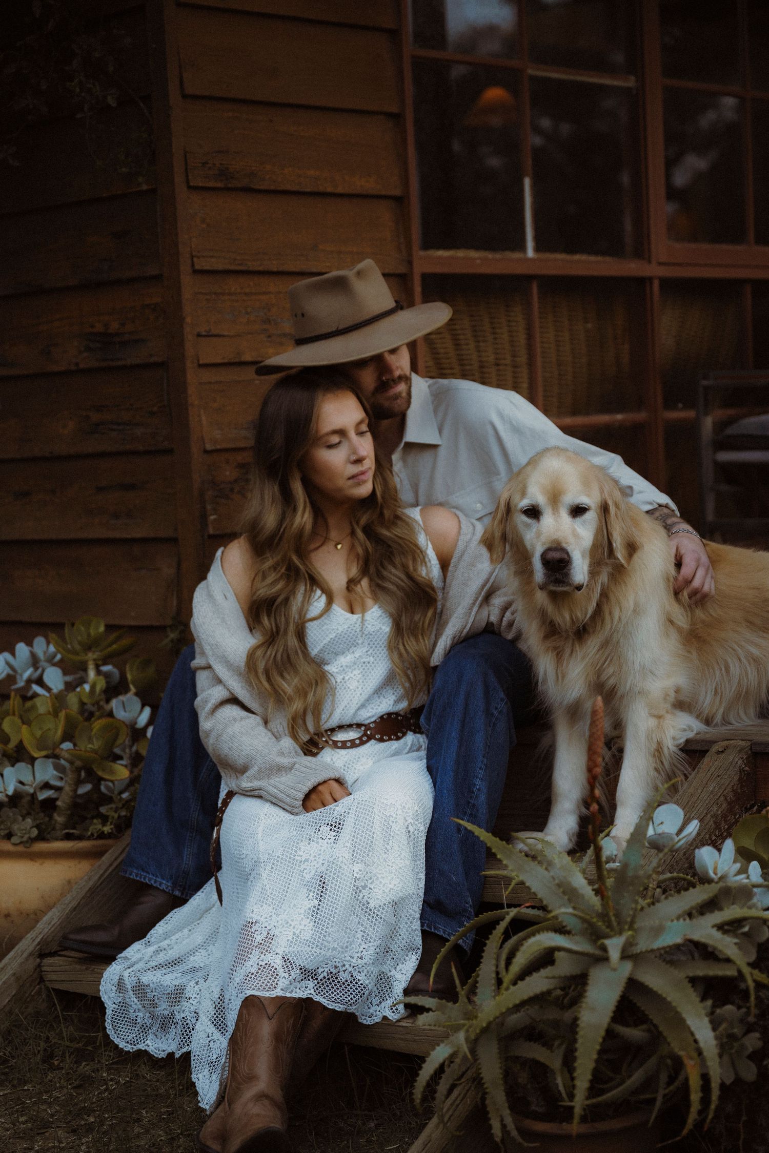Couple sitting together with a Golden Retriever outside a rustic wooden cabin, woman in white lace dress and brown boots.