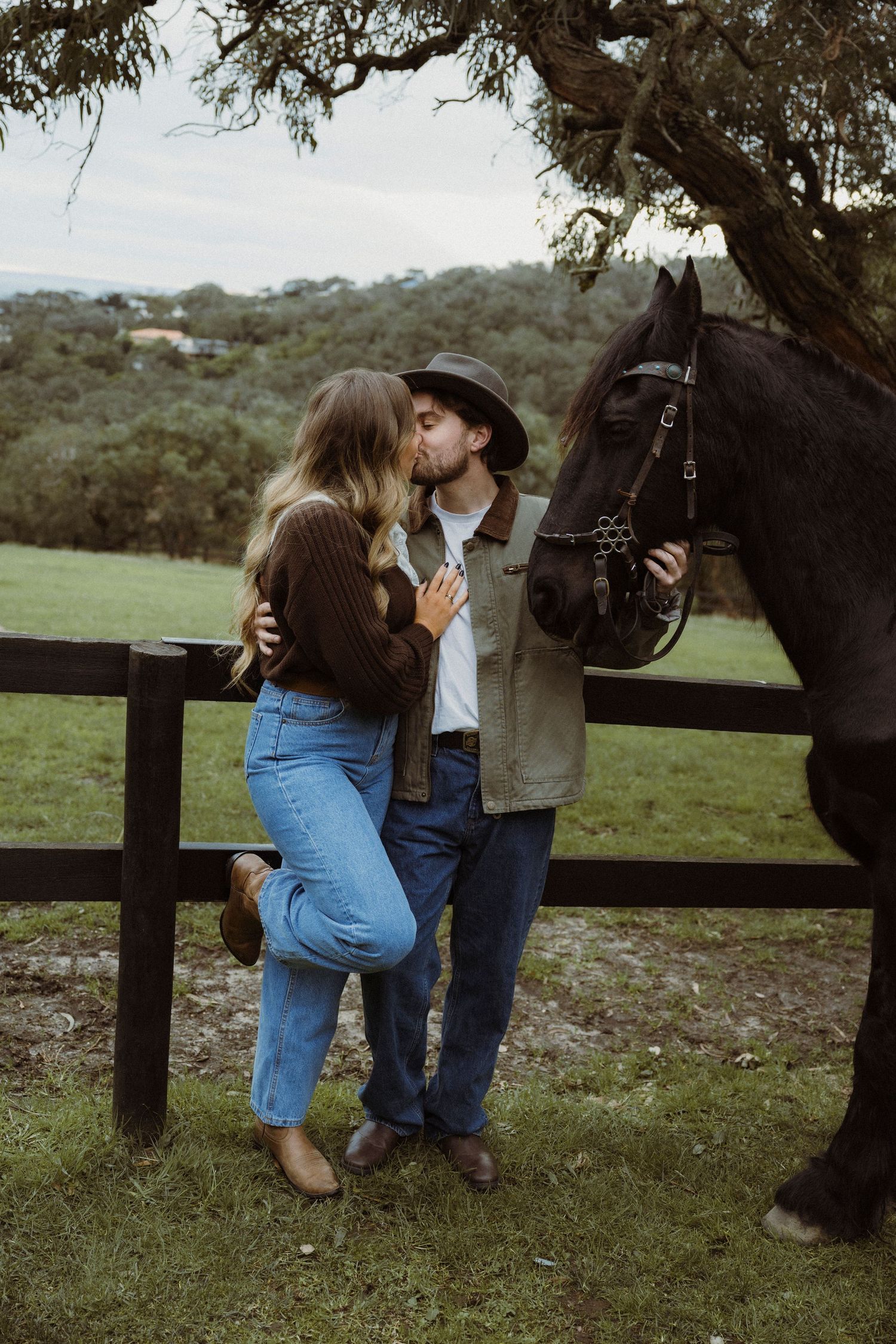 A couple shares a romantic kiss beside a dark horse and wooden fence on a lush green farm.