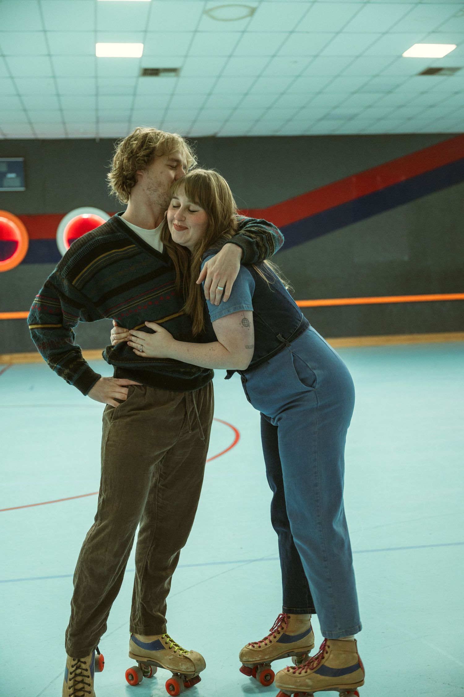Couple wearing roller skates embrace affectionately inside a retro roller skating rink with colorful walls.