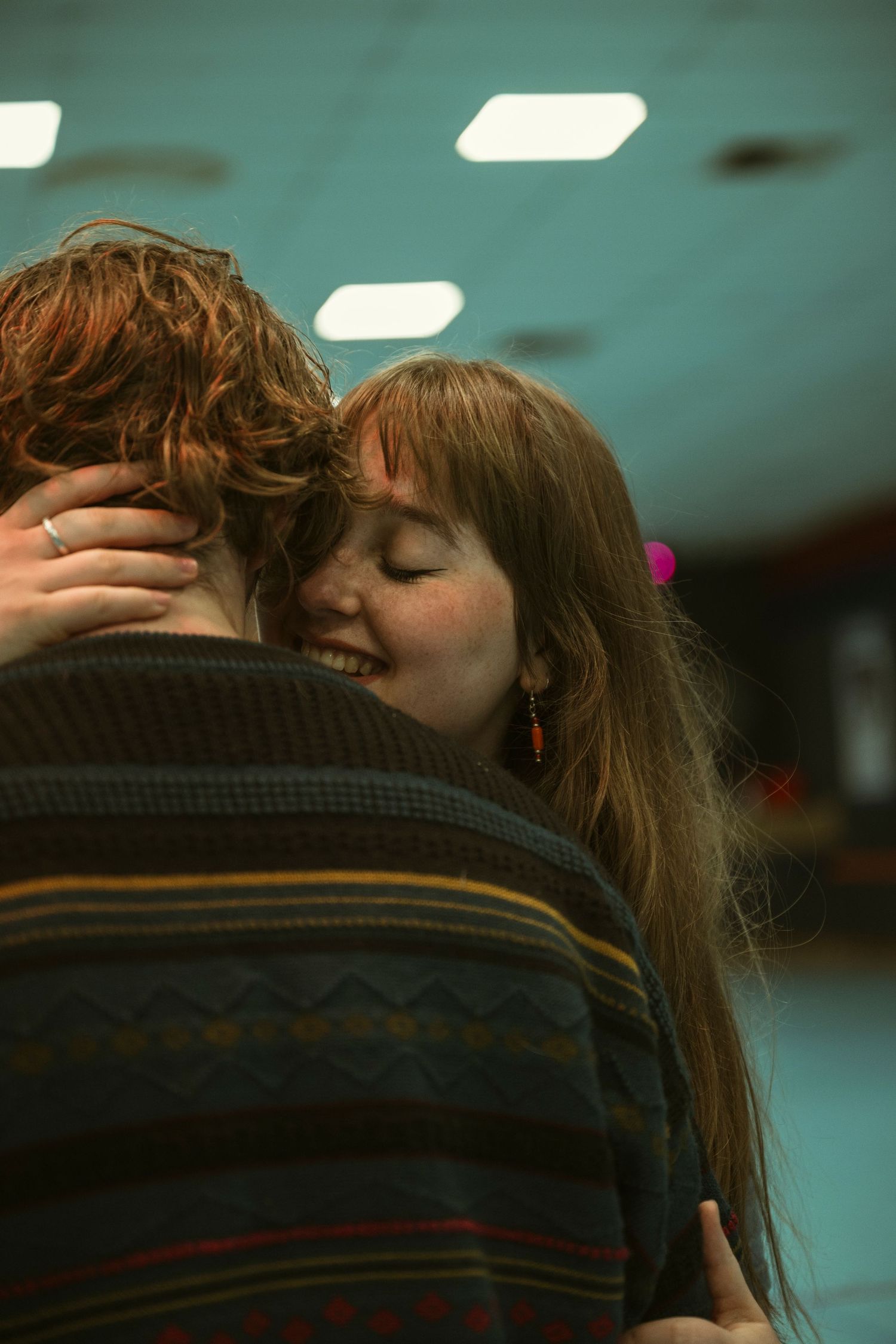 A smiling woman with long hair hugs a man in a striped sweater indoors, eyes closed in a warm embrace.