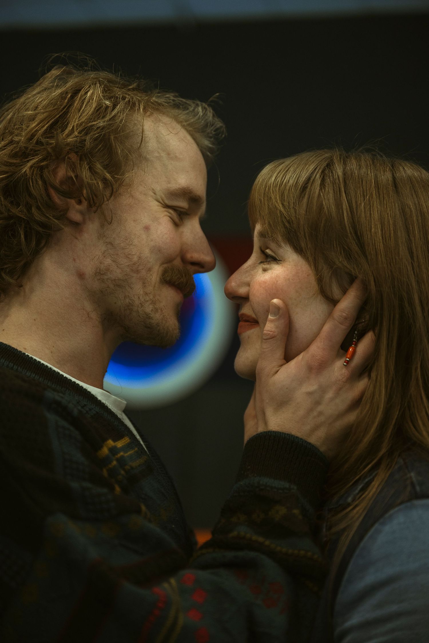 A romantic couple nearly kissing, man tenderly holding womans face near a neon sign in dim lighting.