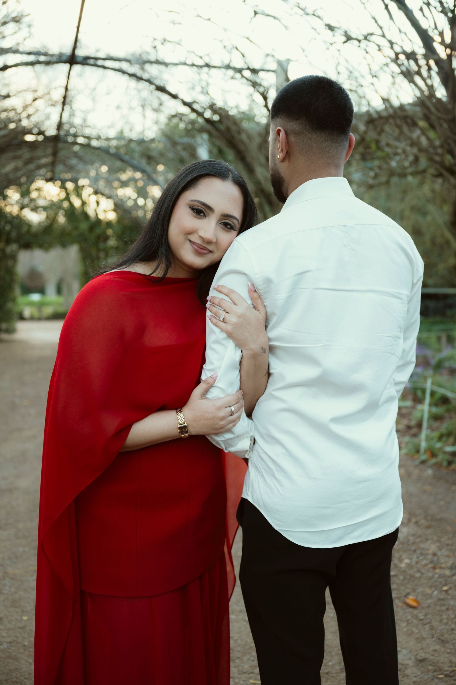 A woman in a red dress embraces a man in a white shirt outdoors in a garden setting.