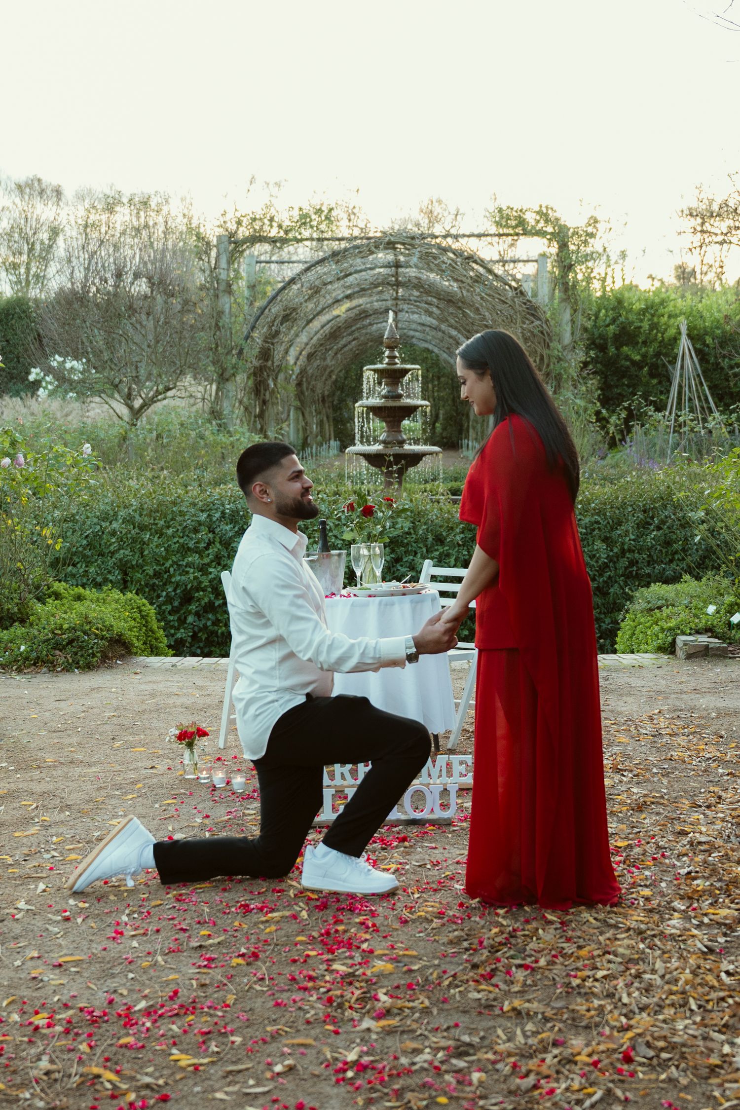 Man proposing on one knee to woman in red dress in a garden with rose petals and fountain backdrop.