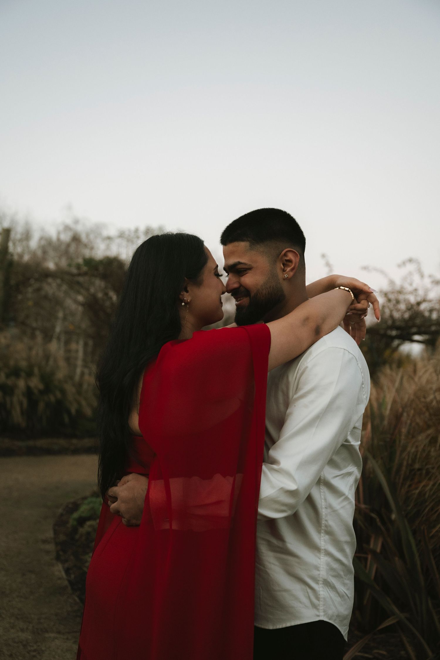 Couple in red and white embracing outdoors, sharing a romantic moment near natural foliage at dusk.