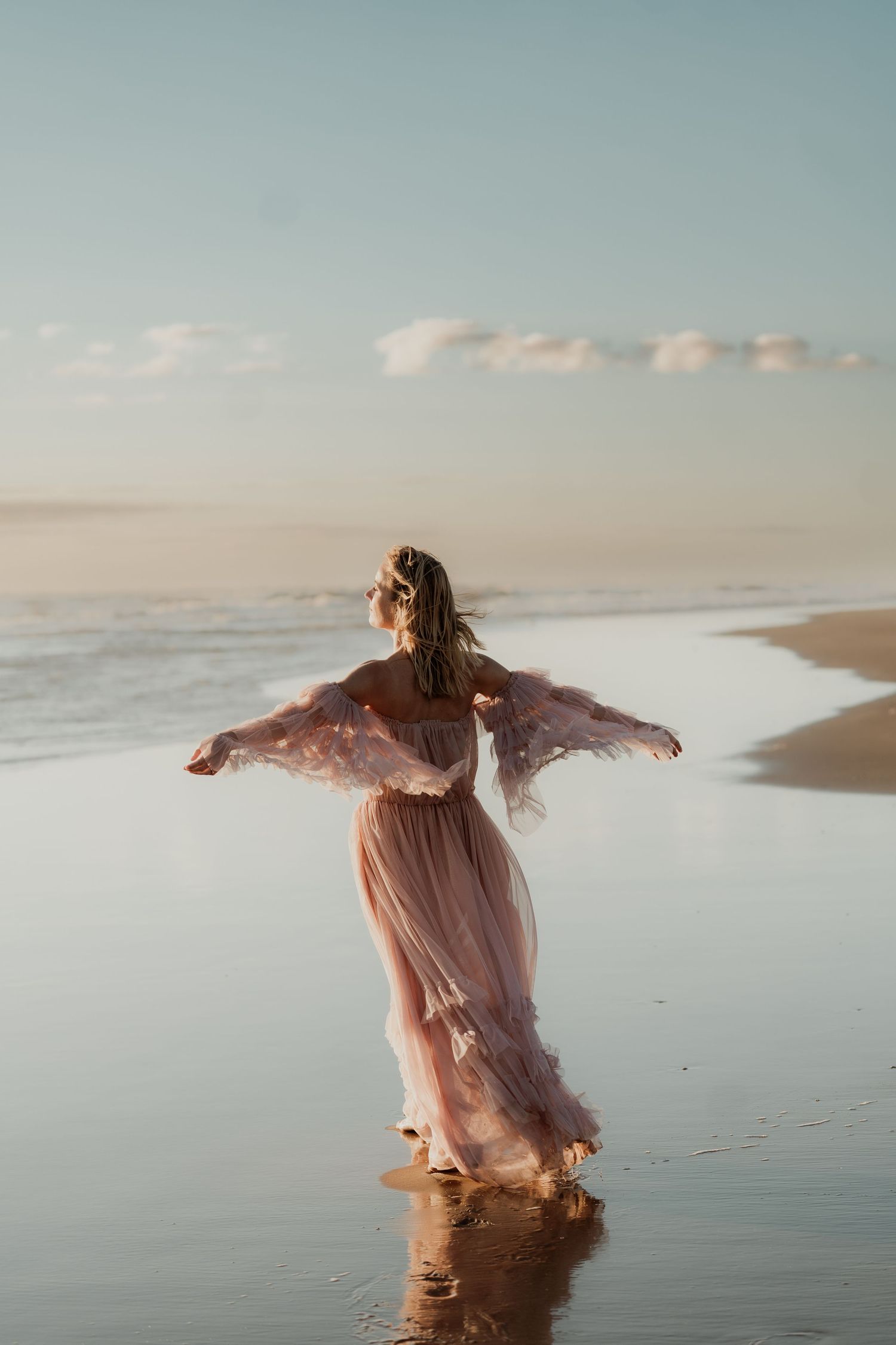 Woman in flowing pink tulle dress walks barefoot on a serene beach at sunset with arms outstretched.