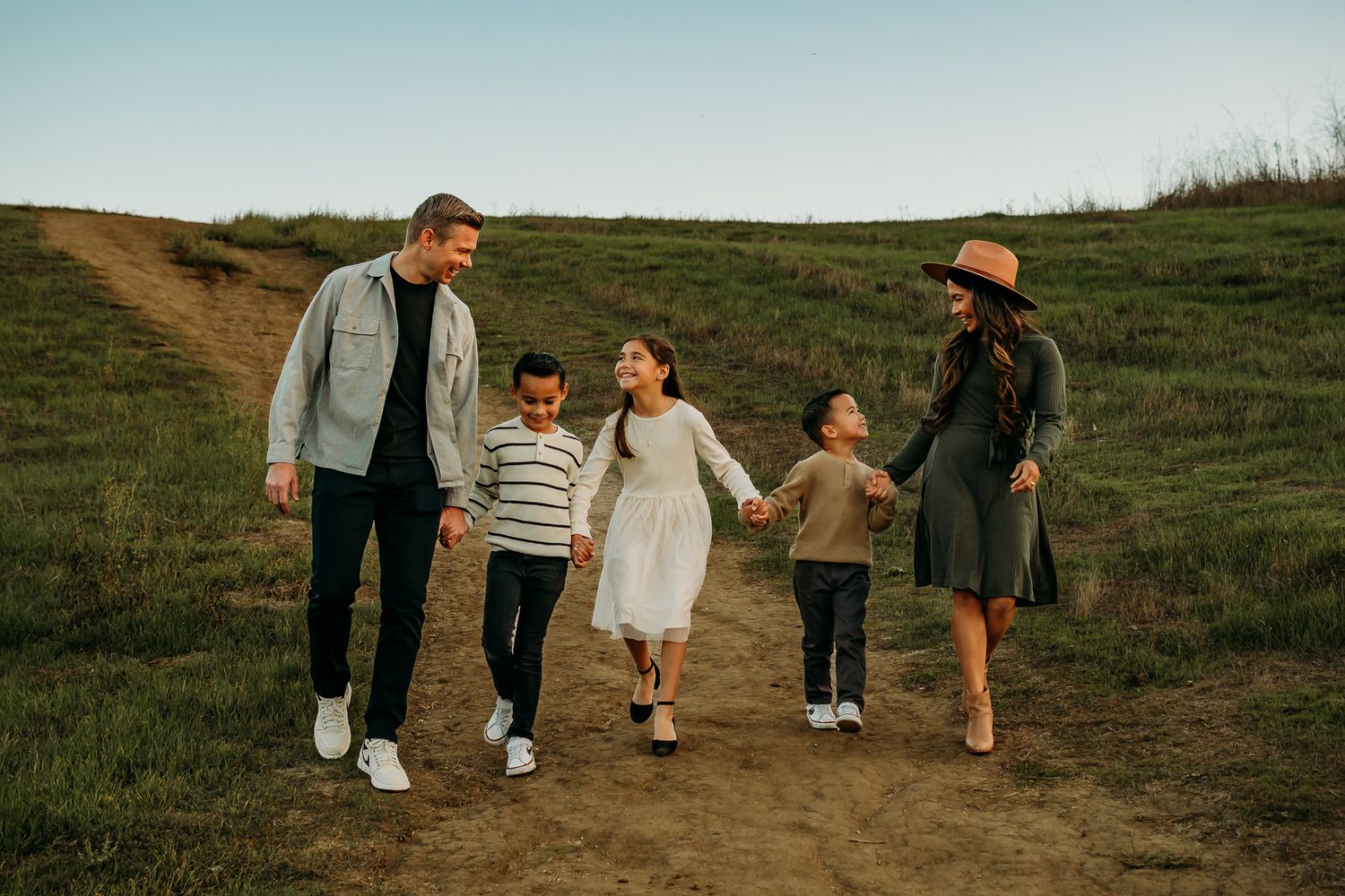 Happy family of five walking together on a scenic green hillside trail, parents holding hands with three children outdoors.