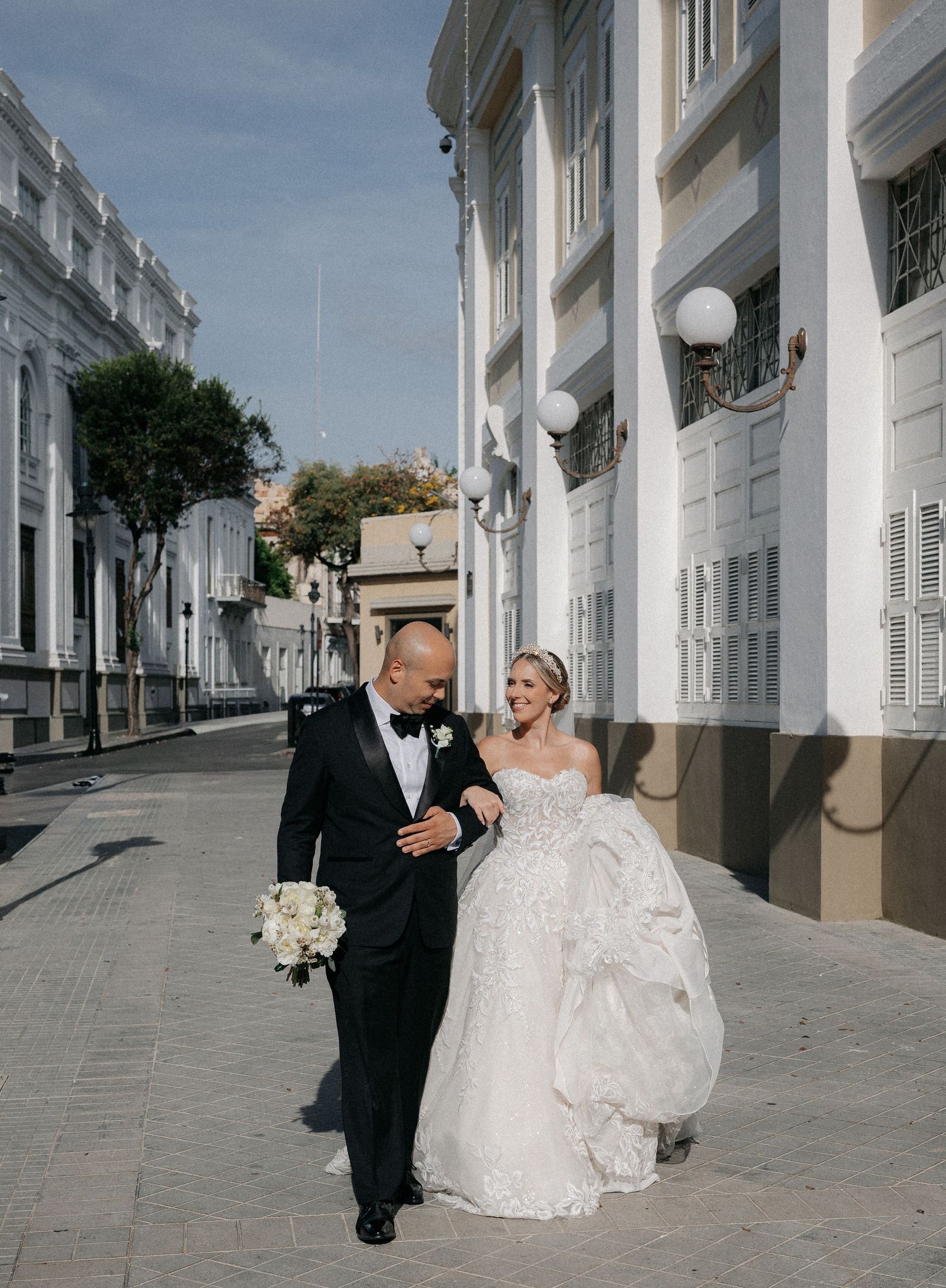 Bride and groom walking arm in arm down a sunlit colonial street, she in a ballgown, he in a tuxedo.