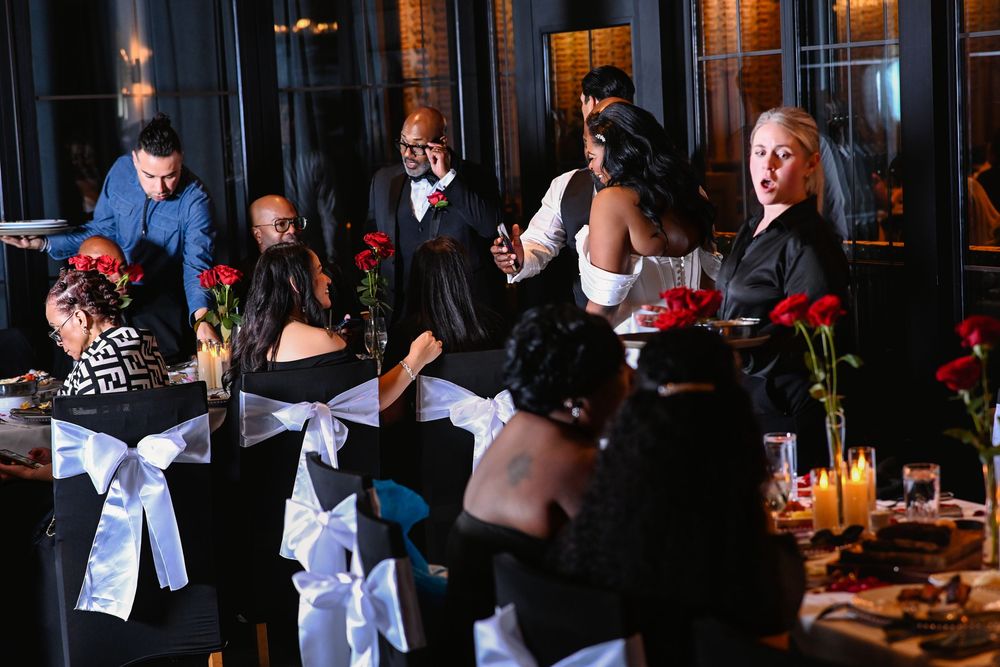 Elegant wedding reception with guests seated at black-draped tables adorned with red roses and candles in a dark venue.