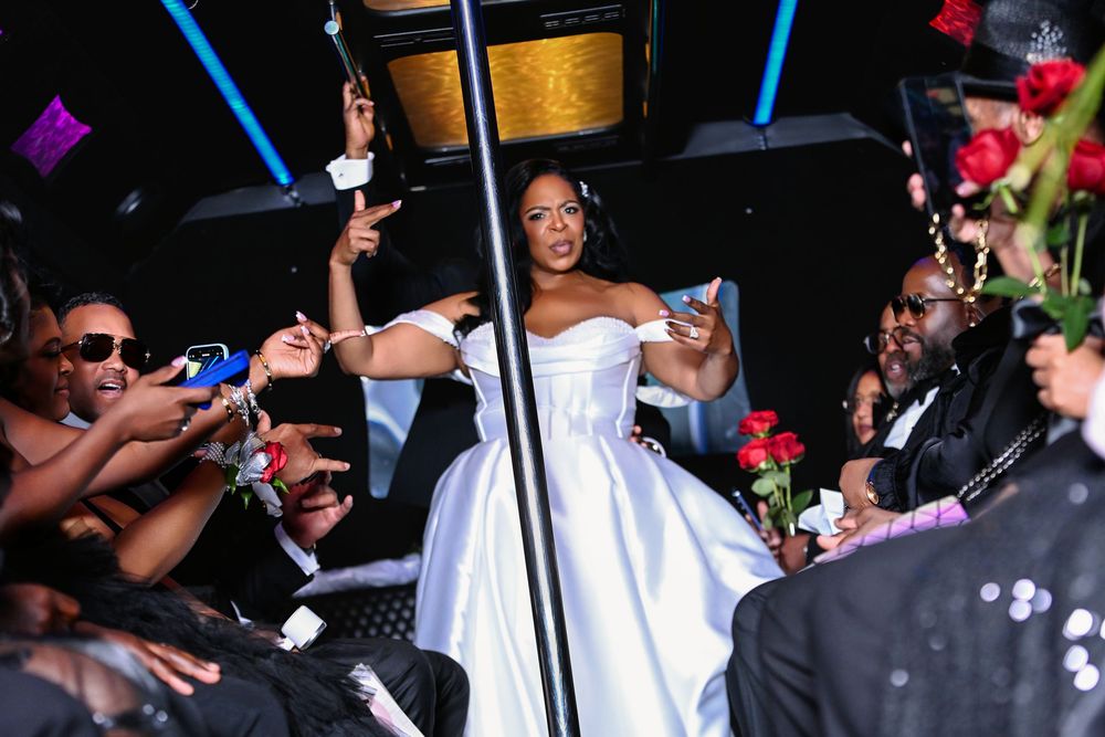 Bride in white off-shoulder gown celebrated by guests holding roses and sparklers at a festive wedding reception.
