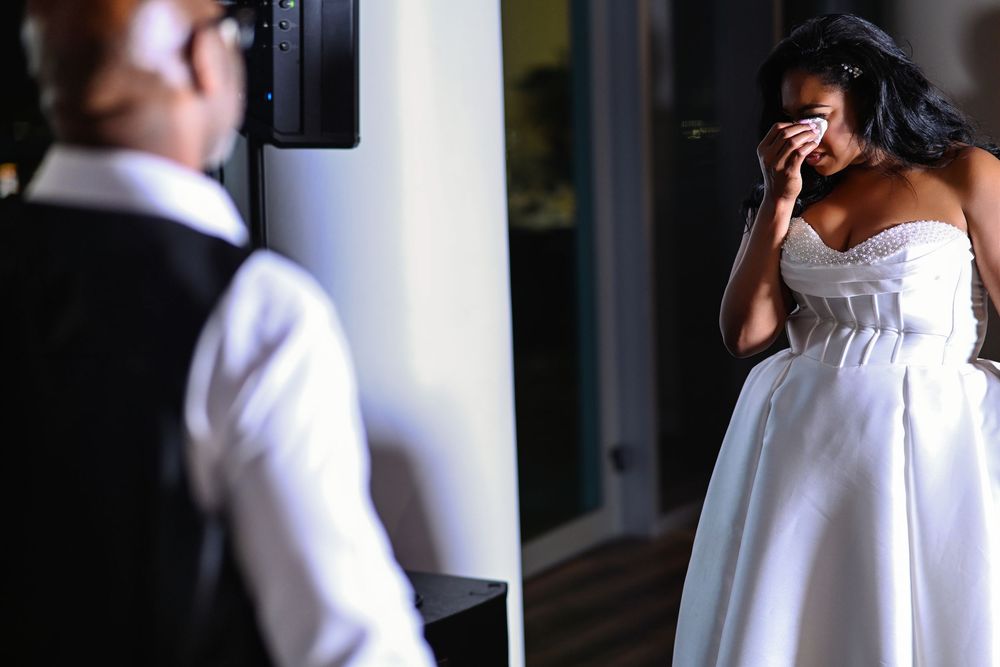 Emotional bride in white strapless gown wiping tears during first look moment with groom at wedding.