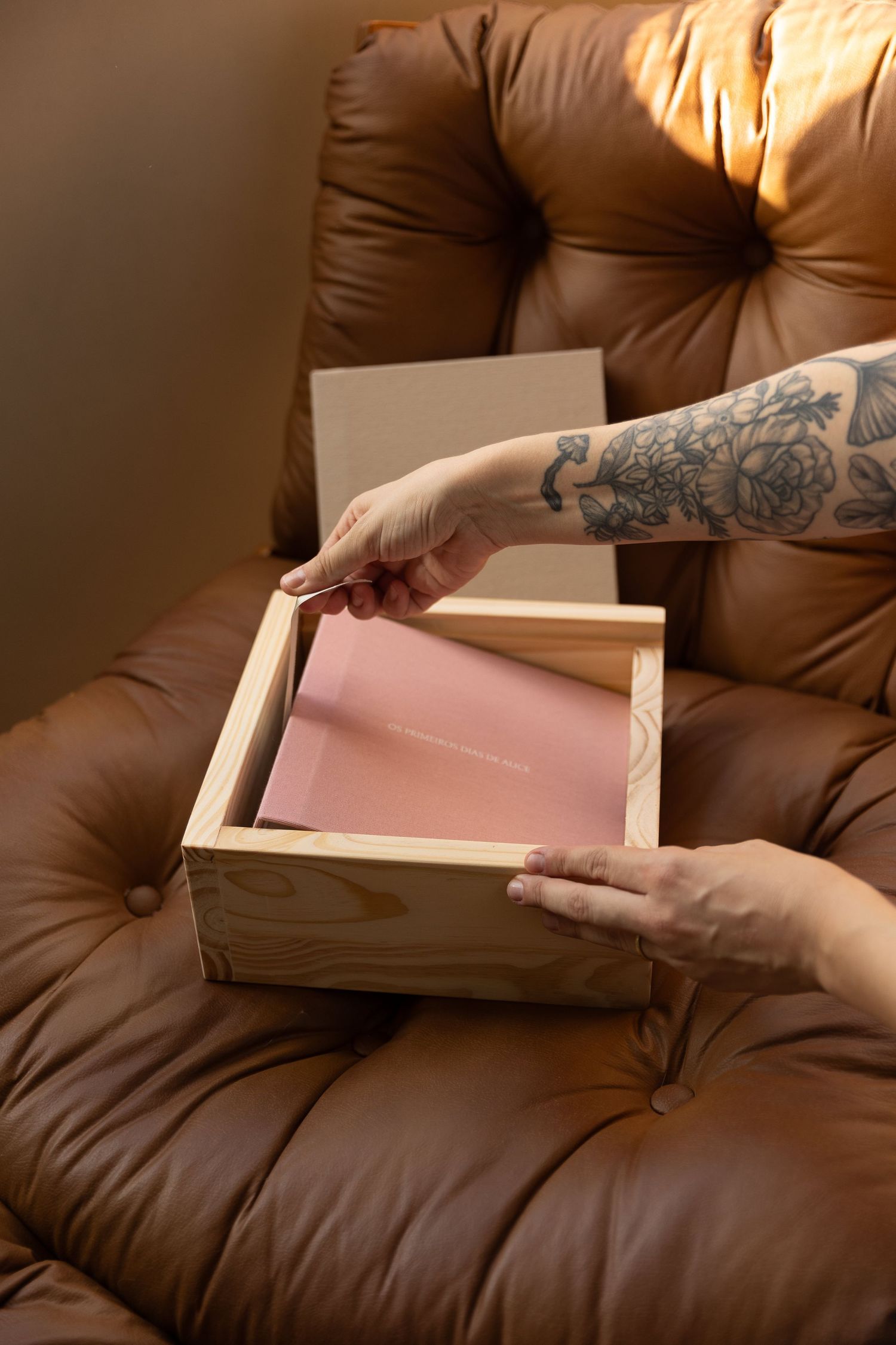 Tattooed hands opening a wooden box with pink lining on a brown leather tufted chair.
