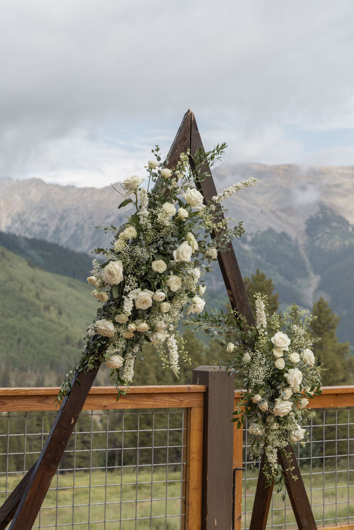 White floral arrangement on wooden pedestal on mountain deck with pine forest backdrop.