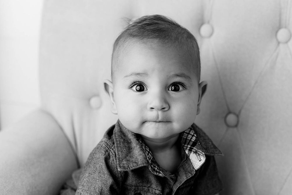 Black and white portrait of an adorable baby boy with big eyes sitting in a tufted chair.