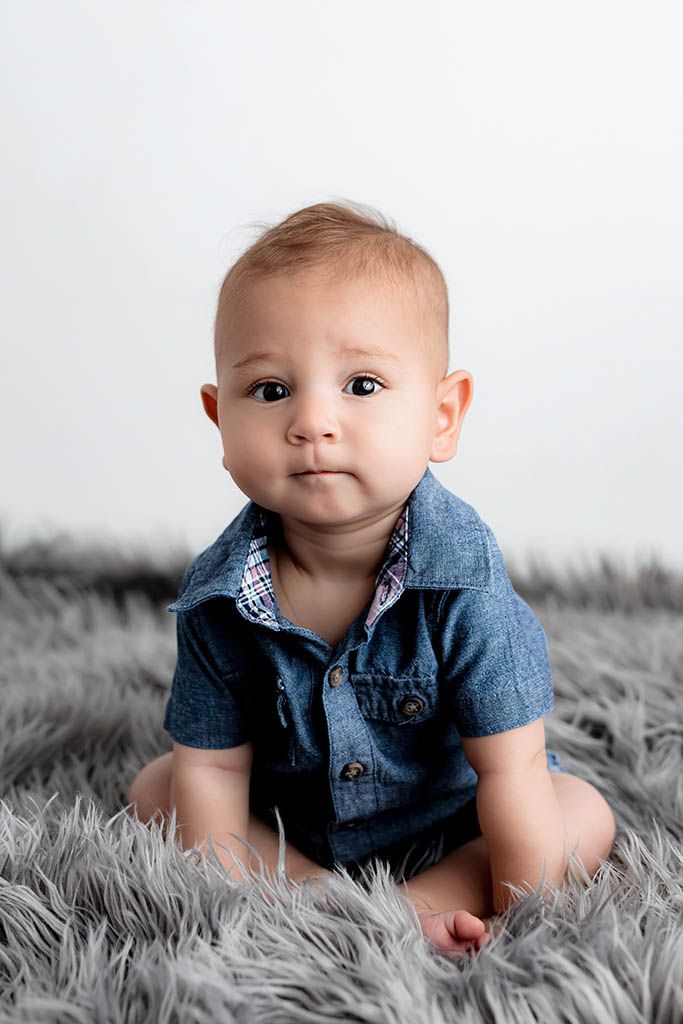Adorable baby boy in denim shirt sitting on gray fluffy rug, looking at camera with big brown eyes.
