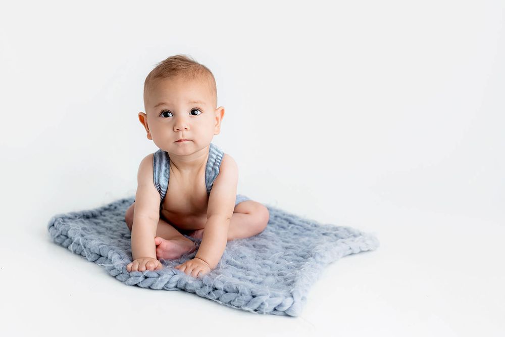 Cute baby sitting on a blue knit blanket against a white background, wearing a light blue romper.