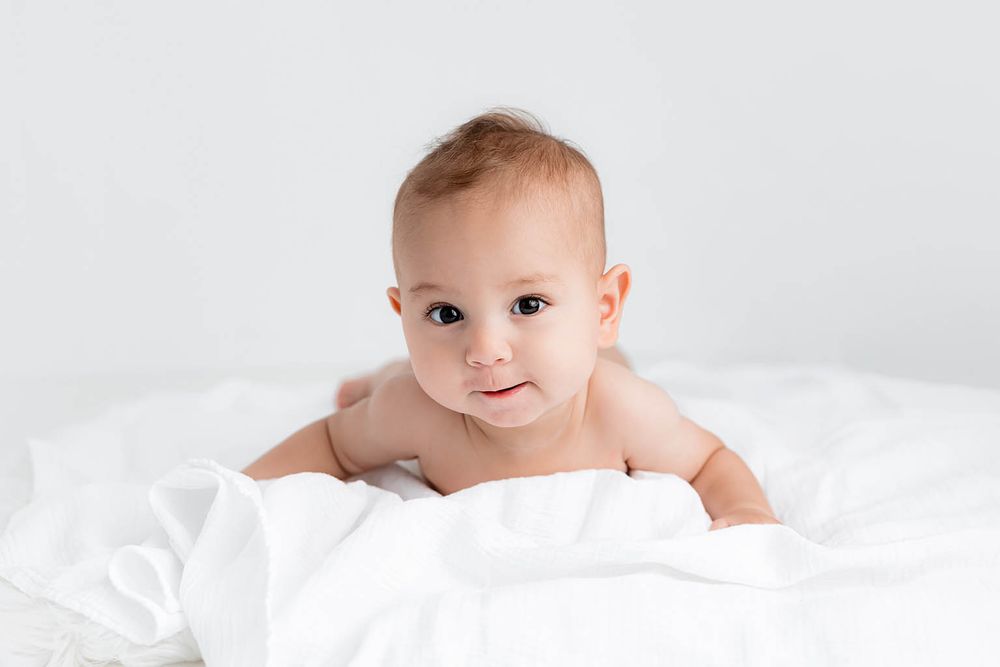 Cute baby lying on white bed covered with soft white blanket, looking at camera with bright eyes.