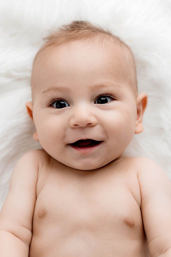 Smiling baby lying on white fluffy blanket with bright eyes and chubby cheeks.