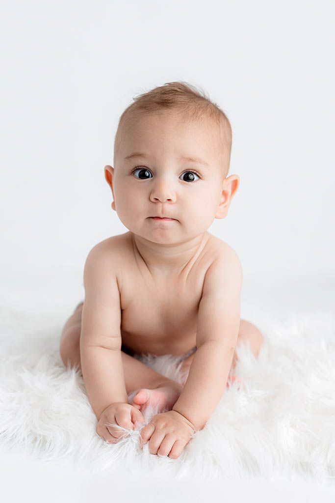 Cute baby sitting on white fur rug with big brown eyes gazing at camera.