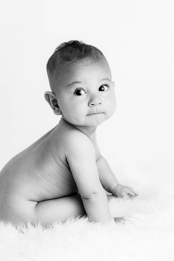 Black and white portrait of a curious baby sitting on a fluffy white rug, looking up with wide eyes.