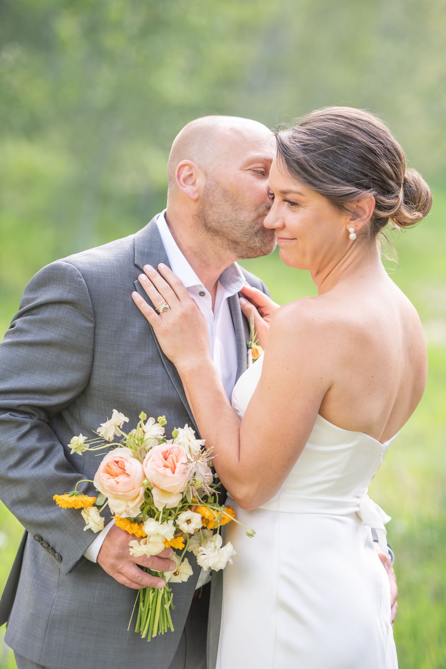 Groom kisses bride on cheek as she holds a peach and yellow floral bouquet outdoors for an elopement in Beaver Creek Colorado