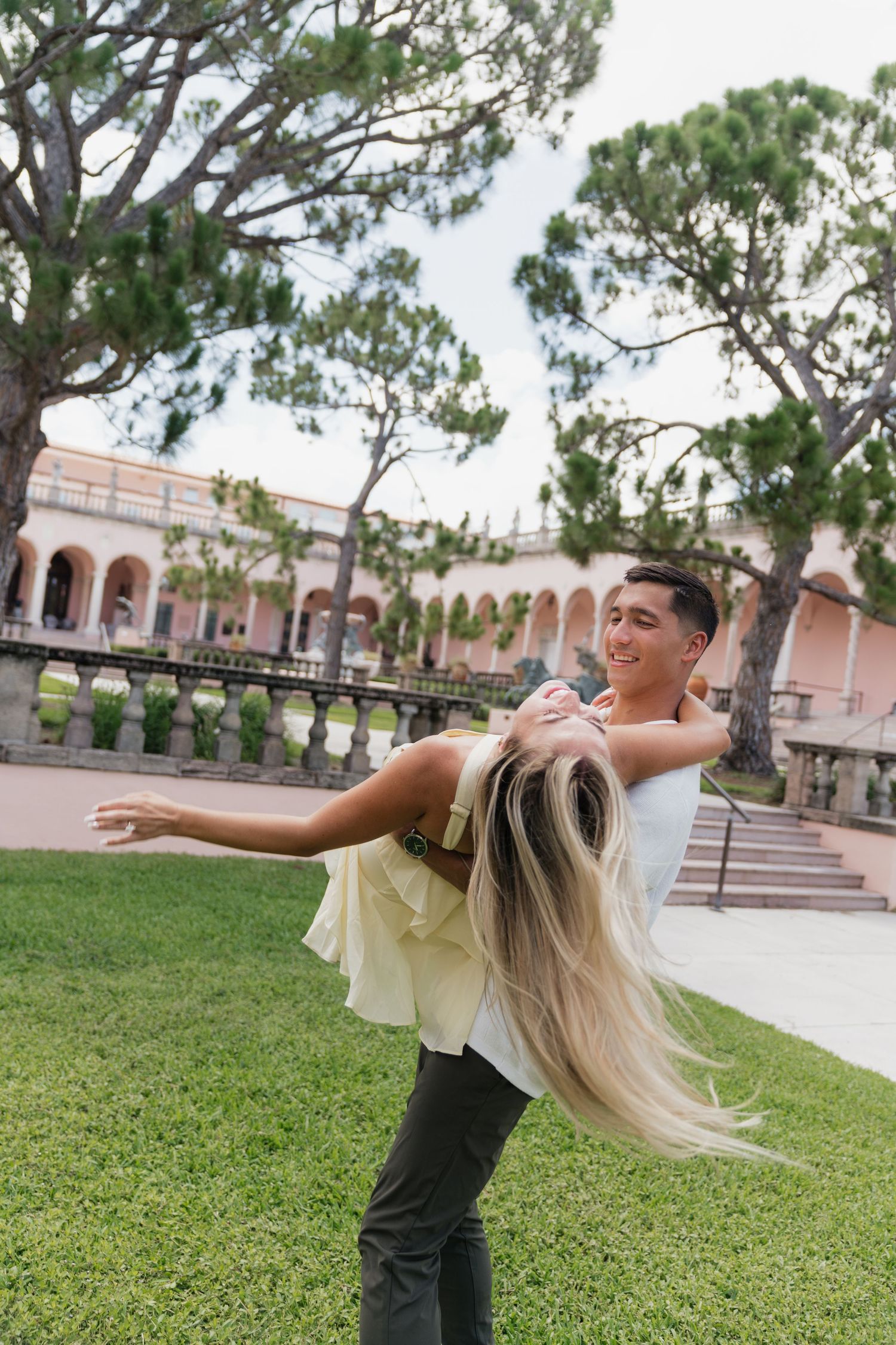 A young man joyfully lifts a blonde woman outdoors near a historic building with lush green lawn.